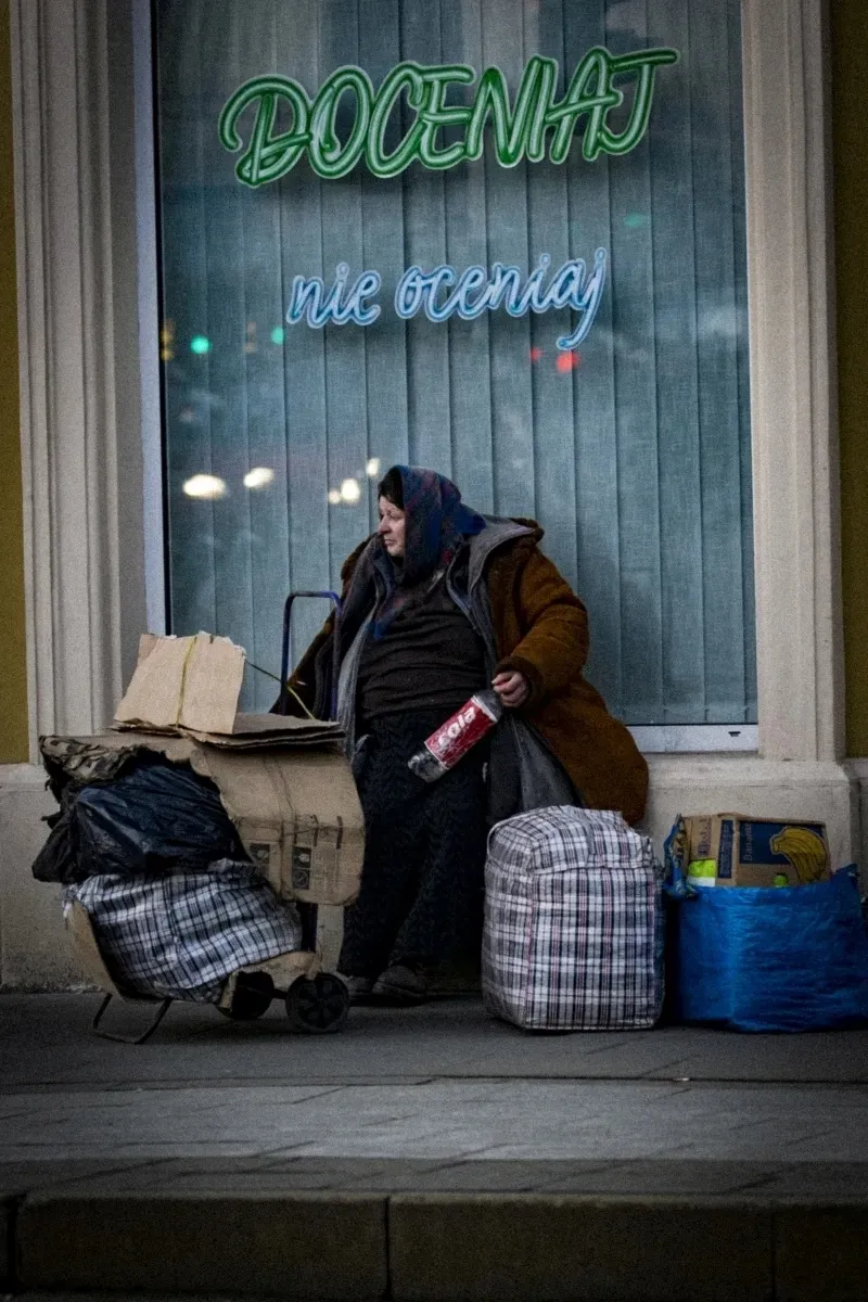 Woman sitting beside bags and a shopping trolley beneath a neon sign reading DOCENIAJ nie oceniaj
